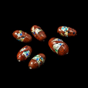 Op de afbeelding: Zes rode, ovale kralen met zilveren, blauwe en groene accenten. De kralen zijn verspreid over een zwarte achtergrond.