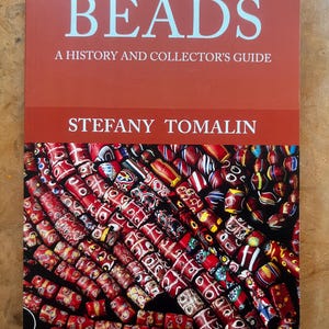 May include: A book titled "BEADS A History and Collector's Guide" by Stefany Tomalin. The cover features a close-up of colorful red, white, and blue beads. The background is a warm, reddish-orange color.