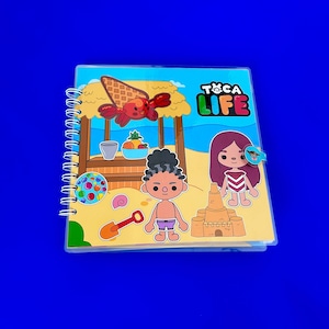 May include: A colorful Toca Life book with a beach scene. The book features a beach hut, a sandcastle, a beach ball, a shovel, a seashell, a crab, and two children. The book has a spiral binding and a clear plastic cover.