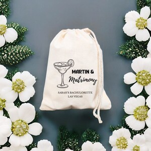 May include: A white cotton drawstring bag with black text that reads "Martin & Matrimony" and "Sarah's Bachelorette Las Vegas". A black and white illustration of a martini glass with a lime wedge is above the text.