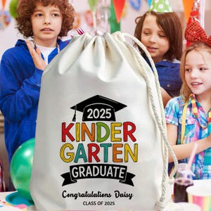 May include: A white drawstring bag with the text "2025 KINDERGARTEN GRADUATE" in colorful letters and a graduation cap graphic. The bag also includes the text "Congratulations Daisy" and "CLASS OF 2025".