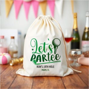 Peut inclure: Un sac en coton crème avec cordon de serrage, orné du texte vert "Let's PARTEE" et d'un motif de balle et de club de golf. Le texte "RON'S 19TH HOLE MIAMI, FL" est imprimé en dessous. Le sac est posé sur une surface en bois.