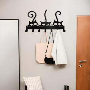 Puede incluir: Perchero de pared de metal negro con cuatro ganchos en forma de gatos. El perchero tiene una base de metal negro y está montado en una pared blanca. Hay dos bolsos y una chaqueta colgados de los ganchos.