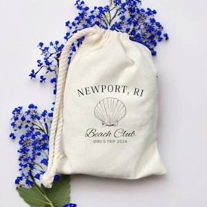 Könnte beinhalten: Eine weiße Kordeltasche mit dem Aufdruck "NEWPORT, RI" und "Beach Club". Die Tasche zeigt außerdem eine Muschelgrafik und den Text "Girl's Trip 2024".