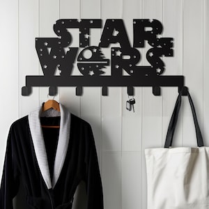 May include: Black metal wall-mounted coat rack with the words "STAR WARS" in a stylized font. The rack has six hooks and features a small Death Star graphic. A black robe hangs on one hook and a white tote bag hangs on another.