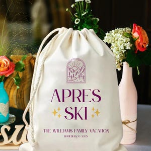 May include: A cream-colored drawstring bag with the words "APRES SKI" in purple, accented with yellow stars. The bag features a mountain and sun graphic and the text "THE WILLIAMS FAMILY VACATION BOULDER CO 2025".