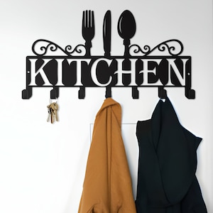 Puede incluir: Un letrero de cocina de metal negro con la palabra "KITCHEN" en letras blancas. El letrero tiene cinco ganchos y presenta un diseño de tenedor, cuchillo y cuchara. Dos chaquetas cuelgan de los ganchos.