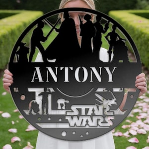 Könnte beinhalten: Runder, schwarzer Metallschild mit Star Wars-Motiven und dem weißen Schriftzug "ANTONY". Das Schild zeigt Silhouetten von Charakteren und das Star Wars-Logo. Das Schild wird von einer Person gehalten.