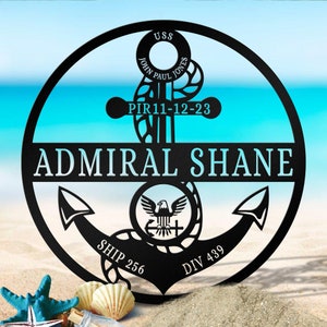 May include: Black metal anchor with a white eagle and the text "USS JOHN PAUL JONES PIR 11-12-23 ADMIRAL SHANE SHIP 256 DIV 439" on a white sandy beach.