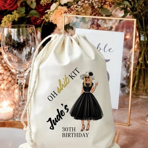 May include: A cream-colored drawstring bag with the text "OH shit KIT" and "Jude's 30th Birthday." The bag features an illustration of a woman in a black dress and sunglasses. The background includes flowers and a wine glass.