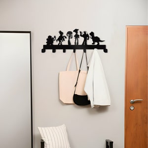 May include: Black metal wall-mounted coat rack featuring a silhouette of characters from the Toy Story movie franchise. The rack has six hooks and is hanging on a white wall. Two bags and a white jacket are hanging on the hooks.