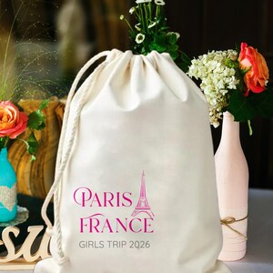 Peut inclure: Un sac à cordon de serrage crème avec les mots "Paris France" et un dessin de la Tour Eiffel en rose. Le texte "GIRLS TRIP 2026" est imprimé en dessous. Le sac a des cordons blancs.