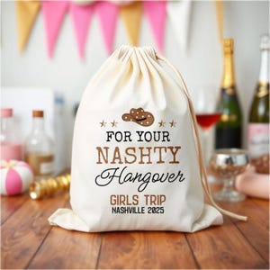 Puede incluir: Una bolsa de cordón crema con el texto "FOR YOUR NASHTY Hangover GIRLS TRIP Nashville 2025" en marrón y negro. La bolsa presenta un sombrero de vaquero y estrellas. El fondo incluye decoraciones de fiesta y botellas.