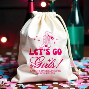 Puede incluir: Bolsa con cordón de color crema con un diseño de bota y sombrero de vaquero rosa. El texto "LET'S GO Girls!" y "SARAH'S BACHELORETTE NASHVILLE 2025" están impresos en rosa. La bolsa está sobre una superficie de madera con confeti.