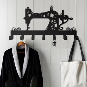 May include: Black metal wall-mounted coat rack shaped like a sewing machine with decorative floral details. The rack has six hooks and is mounted on a white wall. A black robe and a white tote bag are hanging on the hooks.