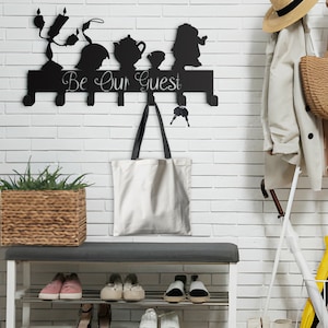 May include: Black metal wall-mounted coat rack with six hooks. The rack features a silhouette of characters from the Disney movie Beauty and the Beast, including Belle, the Beast, and Mrs. Potts. The text "Be Our Guest" is printed on the rack. A white tote bag hangs from one of the hooks.
