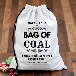Op de afbeelding: Witte trekzak met zwarte tekst die "North Pole Bag of Coal Santa Claus Approved Granted Fresh 100% All Natural" luidt.