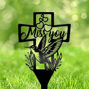 May include: Black metal garden stake featuring a cross design with the words "Miss You" and a hummingbird. The hummingbird is surrounded by leaves and the cross has hearts at the top. The stake is designed to be placed in the ground.