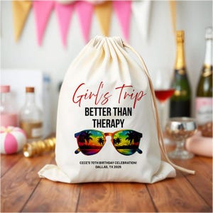May include: A cream-colored drawstring bag with the text "Girl's Trip BETTER THAN THERAPY" in red and black. The design includes sunglasses with a tropical sunset scene. The bag also includes the text "CECE'S 70TH BIRTHDAY CELEBRATION! DALLAS, TX 2025".