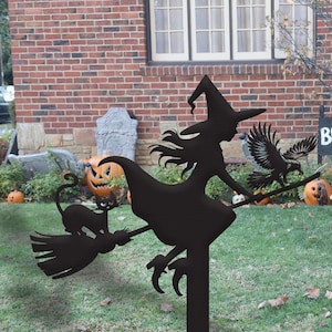 May include: Black metal Halloween yard decoration featuring a witch on a broomstick with a black cat and an eagle. The witch wears a pointed hat and flies past carved pumpkins and a tombstone. The background includes a brick house and green grass.