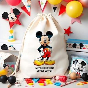 May include: A cream-colored drawstring bag featuring a large Mickey Mouse graphic, with the text "HAPPY 3RD BIRTHDAY GEORGE COOPER". The image includes party decorations like balloons, stars, and a party hat, all in a festive setting.