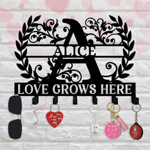 May include: Black metal wall-mounted key holder with the name "ALICE" and the text "LOVE GROWS HERE". The key holder has decorative floral designs and multiple hooks for hanging keys, sunglasses, and other items.