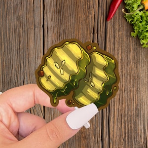 May include: A sticker of two cartoon pickle slices, one green and one yellow, with a dripping effect. The sticker is on a wooden surface.