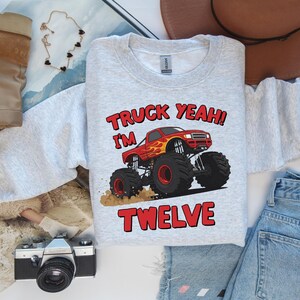 May include: A light gray sweatshirt with the text "TRUCK YEAH! I'M TWELVE" and a graphic of a red monster truck with flames. The sweatshirt is surrounded by a brown hat, sunglasses, jeans, and boots.