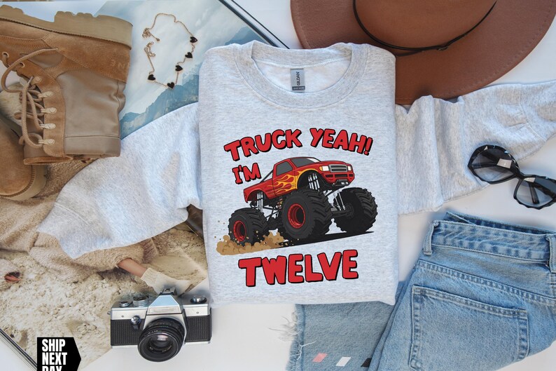 May include: A light gray sweatshirt with the text "TRUCK YEAH! I'M TWELVE" and a graphic of a red monster truck with flames. The sweatshirt is surrounded by a brown hat, sunglasses, jeans, and boots.