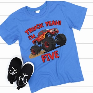 May include: A blue t-shirt with a monster truck graphic and the text "TRUCK YEAH! I'M FIVE" in red. Black and white sneakers are next to the shirt. The shirt features a red monster truck with flames.