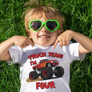 May include: A child wearing a white t-shirt with a red monster truck graphic and the text "TRUCK YEAH! I'M FOUR". The child is wearing green heart-shaped sunglasses and is lying on green grass.