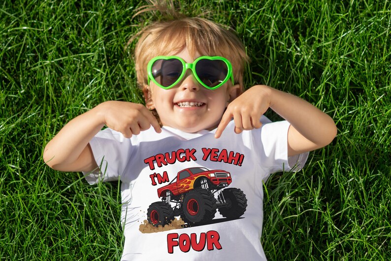 May include: A child wearing a white t-shirt with a red monster truck graphic and the text "TRUCK YEAH! I'M FOUR". The child is wearing green heart-shaped sunglasses and is lying on green grass.