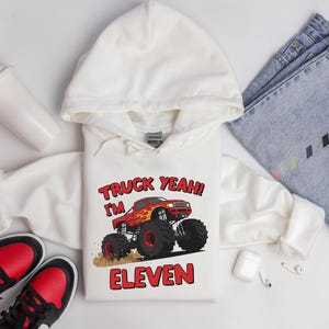 May include: White hooded sweatshirt with a red monster truck graphic and the text "TRUCK YEAH! I'M ELEVEN". The sweatshirt is paired with blue jeans, red and black sneakers, and a white travel mug.