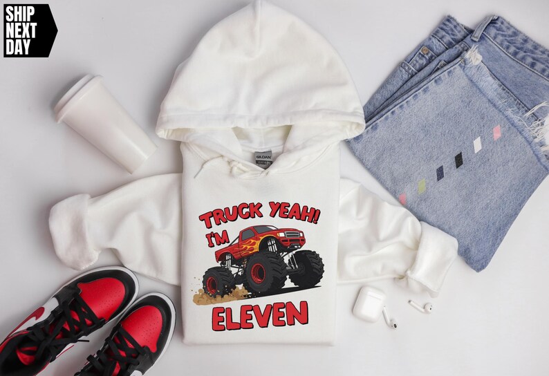 May include: White hooded sweatshirt with a red monster truck graphic and the text "TRUCK YEAH! I'M ELEVEN". The sweatshirt is paired with blue jeans, red and black sneakers, and a white travel mug.
