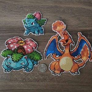 Puede incluir: Tres personajes de Pokemon bordados en punto de cruz: un Bulbasaur azul, un Venusaur azul con una flor rosa y un Charizard naranja con alas azules.