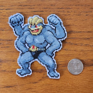 以下が含まれることがあります： ベルトと黄色いモホークを持つ、青と黄色の刺繍されたキャラクター。キャラクターは腕を上げて立っています。刺繍は白い背景です。