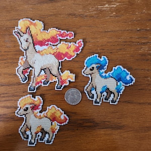 May include: Three cross-stitch patches of stylized horse-like creatures with flame-like manes and tails. One patch has a fiery orange and yellow mane, while another has a blue mane. A quarter coin is next to the patches.