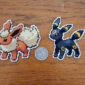 Puede incluir: Dos personajes de Pokemon bordados, un Eevee marrón y blanco y un Umbreon negro y amarillo, se muestran sobre una superficie de madera.