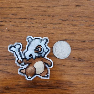 Puede incluir: Un pequeño punto de cruz marrón y blanco de un personaje de Pokemon, posiblemente un Cubone, con un hueso en la mano. El punto de cruz está sobre una superficie de madera junto a una moneda de 25 centavos de dólar estadounidense.