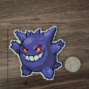 Può includere: Un personaggio di Pokemon Ghost in crochet viola e bianco, di circa 8,25 cm per 8,57 cm, su uno sfondo di legno.