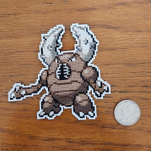 Könnte beinhalten: Eine braune und weiße Kreuzstich-Stickerei eines Monsters mit großen Hörnern und einem segmentierten Körper.