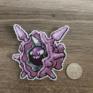 Peut inclure: Un point de croix violet et blanc représentant un personnage de Pokemon. Le personnage est une créature violette et noire avec une corne blanche. Le personnage est entouré d'une bordure blanche.