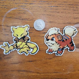 May include: Two embroidered cross-stitch patches featuring Pokemon characters. The first patch is yellow and features a Pokemon named Flareon. The second patch is brown and features a Pokemon named Arcanine.
