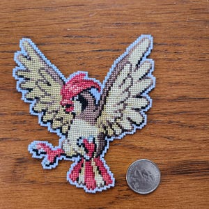 Pode incluir: Ponto de cruz de um pássaro marrom e vermelho com asas brancas, sobre um fundo de madeira. O pássaro é um personagem de Pokemon.