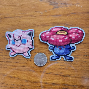 May include: Two embroidered patches, one of a pink Pokemon character and the other of a red and blue mushroom character. The patches are on a wooden surface.