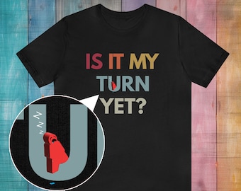 Puede incluir: Camiseta negra con un gráfico de estilo retro que dice "Is it my turn yet?" en colores arcoíris. Una pequeña figura roja está durmiendo en la "U" de la palabra "turn".