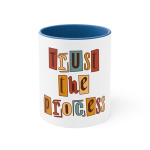 Op de afbeelding: Witte keramische mok met een blauwe rand. De mok heeft de tekst "Trust the Process" erop gedrukt in kleurrijke, retro-achtige letters.