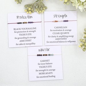May include: Three cards with different gemstone bracelets and descriptions. The first card says "Protection" and lists Black Tourmaline, Tiger's Eye, and Amethyst. The second card says "Strength" and lists Carnelian, Clear Quartz, and Amethyst. The third card says "Warrior" and lists Garnet, Tiger's Eye, and Moss Agate.