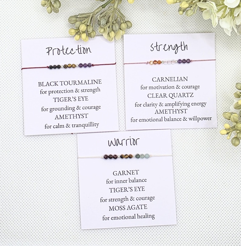 May include: Three cards with different gemstone bracelets and descriptions. The first card says "Protection" and lists Black Tourmaline, Tiger's Eye, and Amethyst. The second card says "Strength" and lists Carnelian, Clear Quartz, and Amethyst. The third card says "Warrior" and lists Garnet, Tiger's Eye, and Moss Agate.