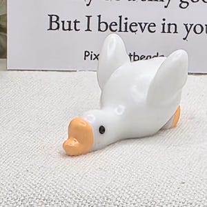 May include: A white ceramic goose figurine with an orange beak and feet. The goose is lying down with its head tilted. Text on a card behind the goose reads, "I may be a silly goose, but I believe in you."